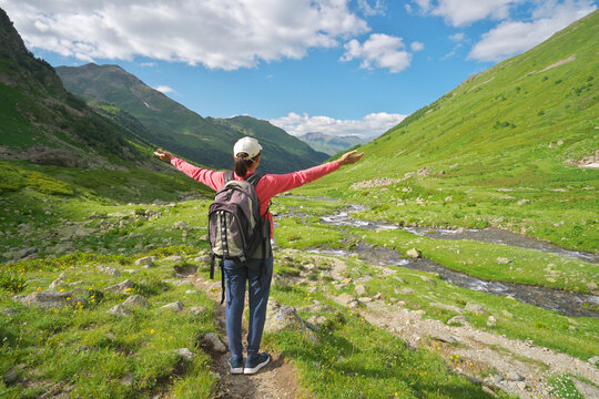 Woman with backpack raises arms standing on a trail in a green mountain valley with a winding river. Active lifestyle image