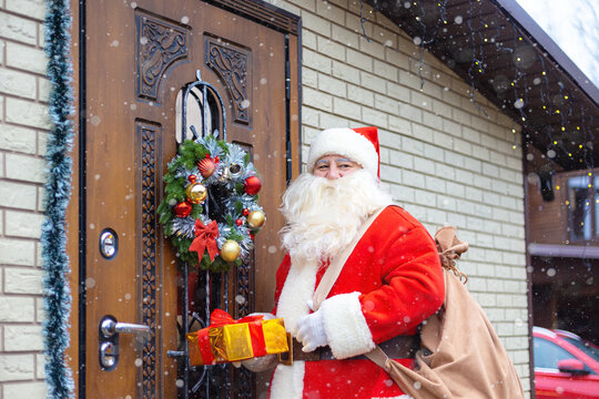 Santa Claus with a gift box stands near a decorated front door.