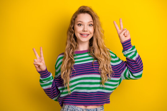 Young woman wearing a striped sweater smiles and shows two peace signs against a bright yellow background