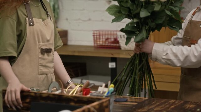 Two young adult Caucasian florists arranging large bouquet of roses in warehouse, tying ribbon and placing flowers on table with tilt down. Clip is ideal for small business marketing