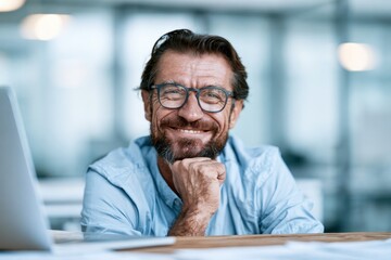 Fototapeta premium Portrait of a mature professional man with glasses and a beard, smiling confidently while sitting at his desk in a bright office
