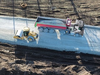 road roller and dump truck are processing gravel for a new road through previously undeveloped rural land  construction site from a bird's-eye view  road construction  environment  development © keBu.Medien