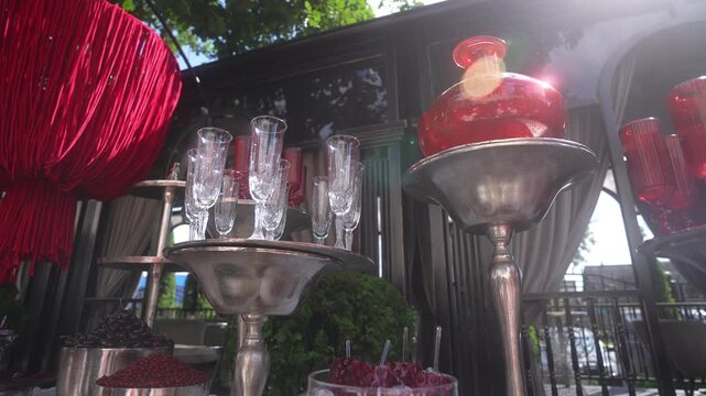 Low-angle outdoor event table display with stacked clear champagne flutes on silver pedestals beside red glass decanter, bright sunlight flare, reflections, elegant festive decor