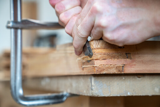 Repairman removes protruding parts of wood patches on old wooden window frame using chisel instrument, experienced carpenter cuts off excess of wood and evens structure of wooden frame at workbench.
