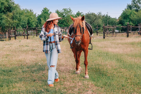 Rustic log fence and open pasture frame a young woman in a felt hat holding the lead rope of a saddled chestnut horse. Rural lifestyle concept.