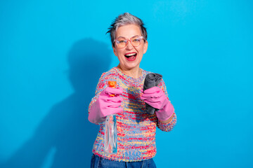 Cheerful senior woman cleaning with spray bottle and cloth wearing pink rubber gloves and colorful...
