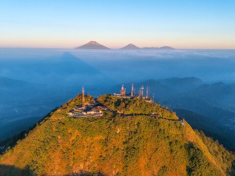 Aerial view of Mount Telomoyo with communication towers illuminated by golden hour light above a sea of clouds in Kecamatan Ngablak, Jawa Tengah, Indonesia.