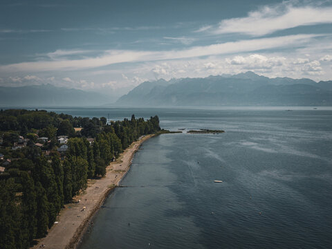 Aerial view of Lake Geneva shoreline with lush trees, a sandy beach, and distant mountains under a blue sky in Morges, Vaud, Switzerland.