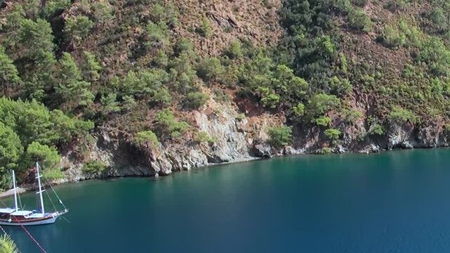 A majestic sailboat anchors in a tranquil Mediterranean bay, embraced by vibrant green forests and rugged cliffs. The water shines a captivating emerald under the clear sky.