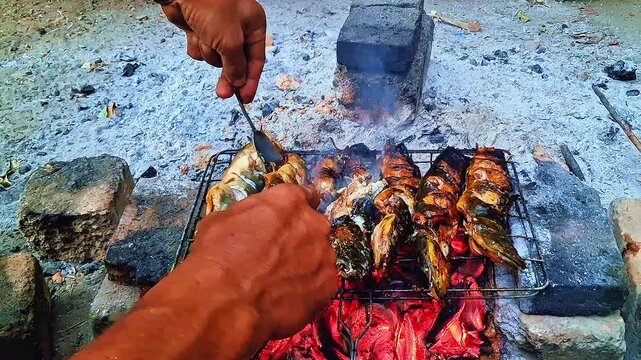 Using a spoon and metal tool to carefully flip and turn whole catfish grilling evenly over a fiery charcoal stove.
