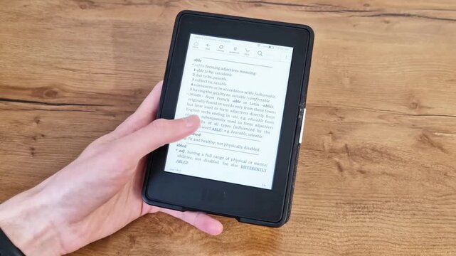Hand holds electronic book reader with open page on wooden table surface