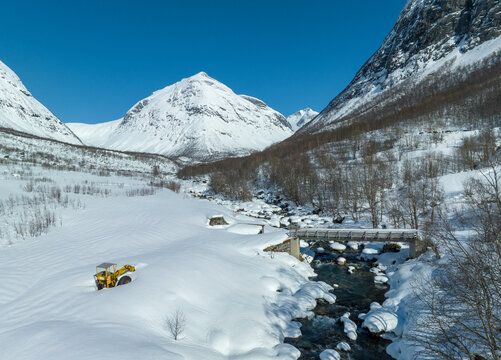 Aerial view of Langdalen valley with a snow-covered landscape, a river, a bridge, and a yellow construction vehicle in the deep snow under a clear blue sky in Fjord, More og Romsdal, Norway.