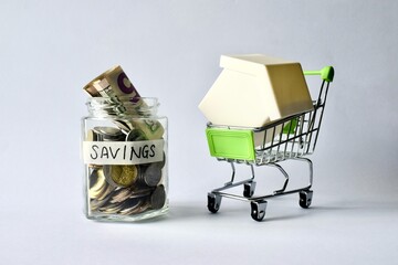 Glass jar labeled SAVINGS next to miniature shopping cart carrying toy house model. © Akram