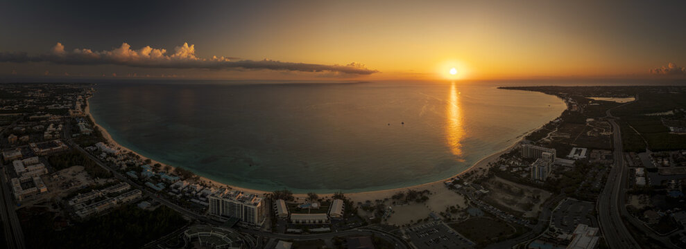Aerial view of Seven Mile Beach at sunset with golden light reflecting on the calm Caribbean Sea, coastal resorts, and lush tropical vegetation in Grand Cayman, Cayman Islands.