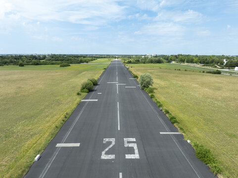 Aerial view of Landebahn Neubiberg, a former airfield runway marked with the number 25, surrounded by lush green fields and trees under a bright blue sky in Unterhaching, Bavaria, Germany.
