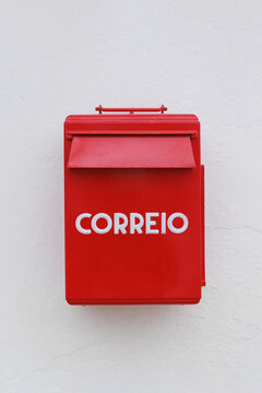 Close-up of a traditional red Portuguese mailbox mounted on a white textured wall, featuring a mail slot, raised lettering, and a clean minimalist composition. Vibrant red color contrasts