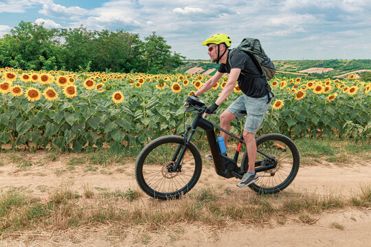 Man in green helmet rides an electric mountain bike along a dirt path beside a vast sunflower field in summer. Adventure concept.