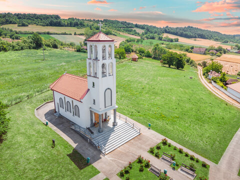 Serbian Orthodox church and bell tower dominating a peaceful rural village with green fields and distant hills under a cloudy sky