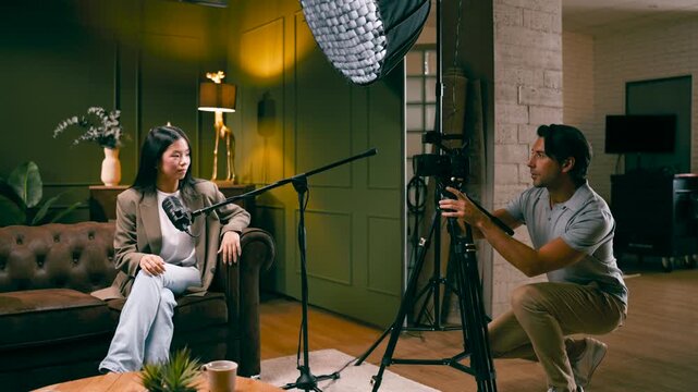 Professional male videographer recording video interview with an asian female influencer sitting on a sofa and talking into a microphone