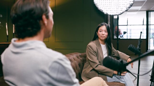 Over the shoulder view of a male host interviewing an asian female guest. Both are sitting on a couch in a studio, talking into microphones