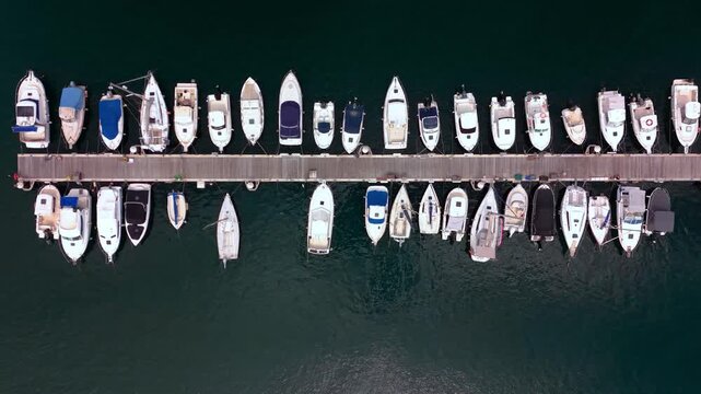 Top down overhead aerial view of recreational motorboats and sailboats moored along a wooden pier in a marina. Nautical luxury travel, maritime transport, and Mediterranean summer harbor concept. High