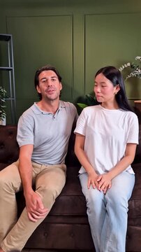 Beautiful couple, an asian woman and a caucasian man, sitting on a sofa while recording a video for social media