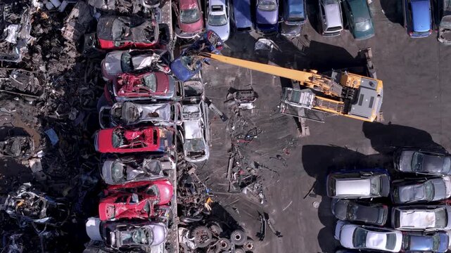 Top-down aerial view of heavy crane handling wrecked cars in scrap yard for industrial recycling and circular economy
