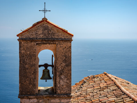 Aerial view of a historic stone church bell tower with a bronze bell and cross, overlooking the blue Mediterranean Sea in Eze, Provence-Alpes-Cote d'Azur, France.