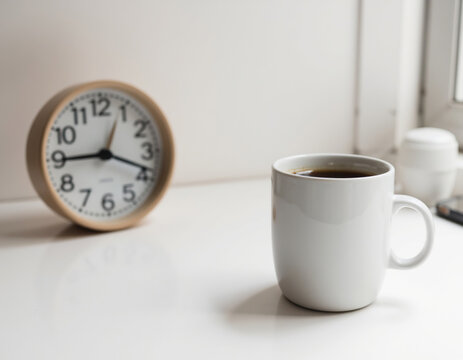 White ceramic coffee mug with black coffee on desk and blurred wooden alarm clock in minimalist workspace setup