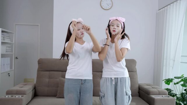 Two young Asian women having fun with a beauty routine, wearing facial sheet masks and pink headbands while sitting on a sofa. Happy friends enjoying a home spa and skincare self-care together.