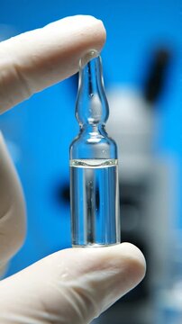 Laboratory technician in white gloves holds a clear glass ampoule filled with liquid, showcasing the ampoule against a blue background with a microscope in view