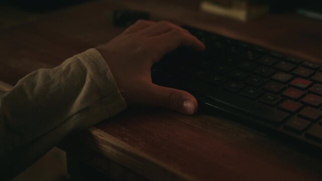 Close-up of a child's hand using a computer keyboard in low light. Technology, gaming, online learning and screen time concept. 4K.