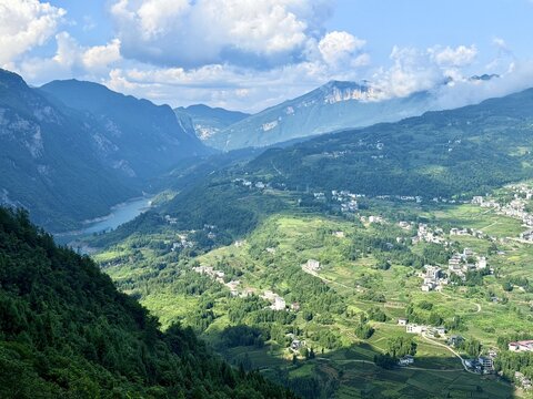 Aerial view of Enshi Grand Canyon featuring lush green mountains, a winding river, and small settlements nestled in the valley under a bright blue sky in Enshi City, Hubei, China.