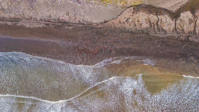 Aerial view of ocean waves washing onto a dark sandy beach covered with scattered plastic debris and waste in Indonesia.