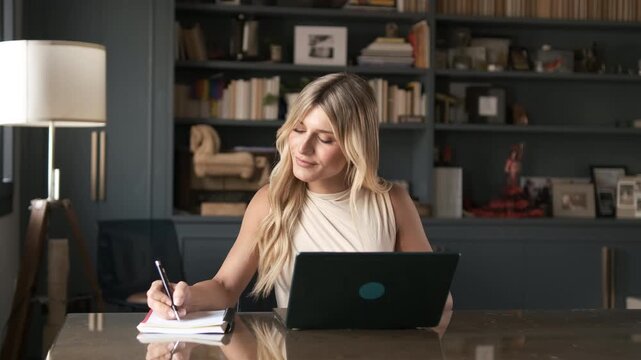 Young transgender freelancer woman writing in diary as she works on laptop at table in home office