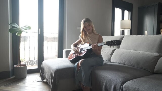 Focused young transgender woman smiling while playing guitar as she sits on sofa in living room at home