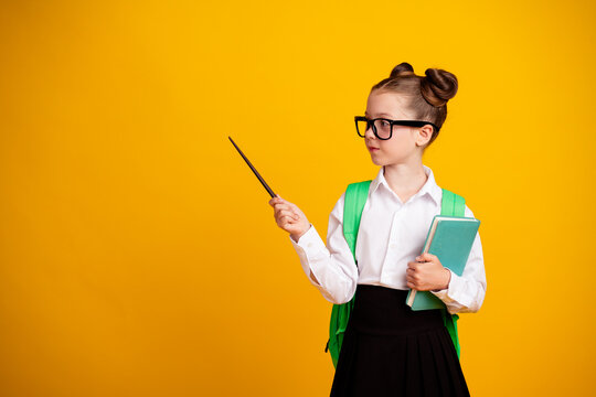 Smart schoolgirl with glasses and green backpack holding a book and pointing with a pointer against a yellow background