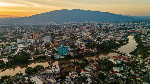 Aerial view of Chiang Mai city skyline featuring the Ping River winding through the urban landscape with lush mountains in the background under a warm golden hour sky, Chiang Mai, Thailand.