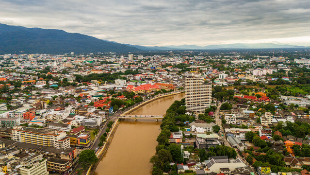 Aerial view of the Ping River flowing through the city of Chiang Mai with a bridge, dense urban buildings, and mountains in the background in Chiang Mai, Thailand.