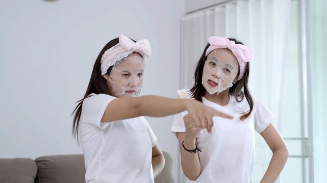 Two young Asian women having fun with a beauty routine, wearing facial sheet masks and pink headbands while sitting on a sofa. Happy friends enjoying a home spa and skincare self-care together.
