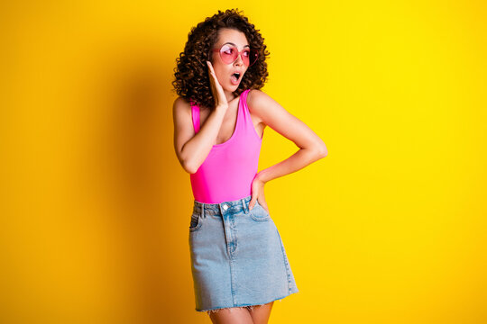 Surprised young woman in pink top and denim skirt posing with heart sunglasses against a bright yellow background, expressive retro fashion portrait conveying fun and playful summer vibes