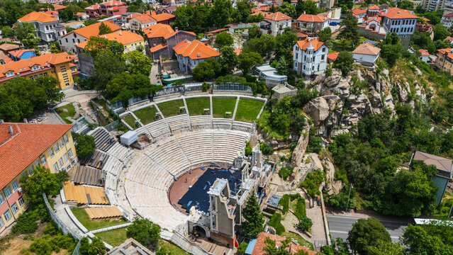 Aerial view of the Ancient Theatre of Philippopolis surrounded by historic buildings, lush greenery, and rocky terrain in Plovdiv, Bulgaria.