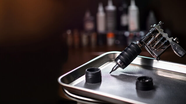 Rotary tattoo machine on metal tray with blurred tattoo studio background banner.