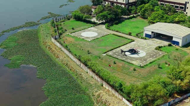 Aerial view of a helicopter parked on a private helipad near a lake with lush greenery and a hangar in India.