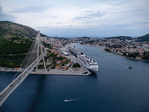 Spectacular aerial view of the Franjo Tuđman Bridge in Dubrovnik, Croatia, straddling a tranquil sea inlet under soft light, framed by rugged coastal peaks and harbor docks.