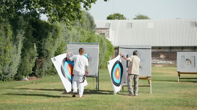 Two archers walking on a green field and replacing old paper targets with new ones. Preparation for a competitive archery tournament
