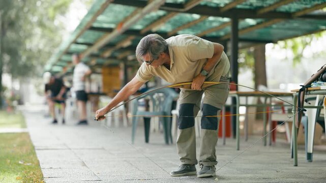Focused senior man carefully stringing his traditional wooden longbow. Outdoor archery range setting for a day of recreational practice