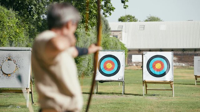 Blurry senior man practicing archery in a field. Concentrated archer holding a bow and aiming at a colorful target during training outdoors
