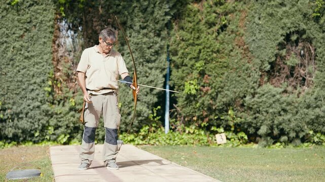 Focused senior man practicing archery on a sunny day. He carefully nocks an arrow, draws the bowstring, aims, and releases the shot