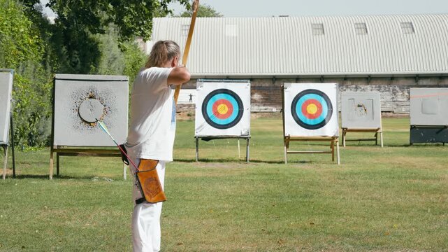 Senior man practicing archery with a longbow, aiming and shooting an arrow at a target on a green field. A moment of focus and skill